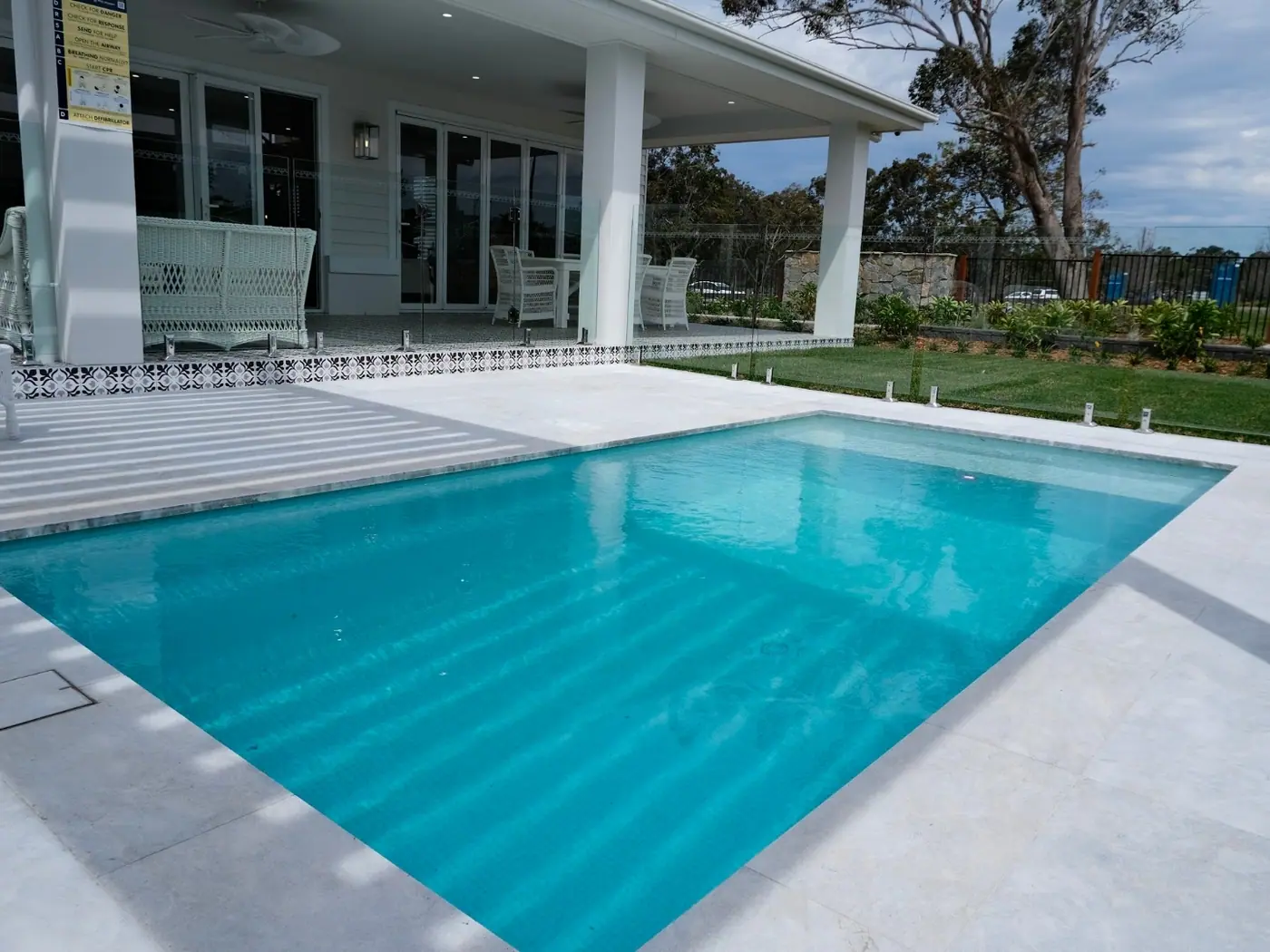 A rectangular plunge pool with pale stone coping directly in front of a covered verandah, with lawn and open paddock fencing beyond.