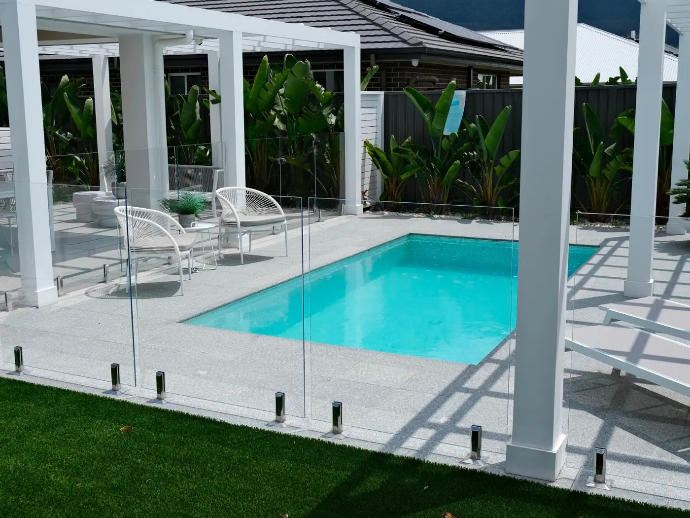 A plunge pool with a wide exposed aggregate surround, white pergola posts either side and glass fencing, with lawn running up to the paving in the foreground.