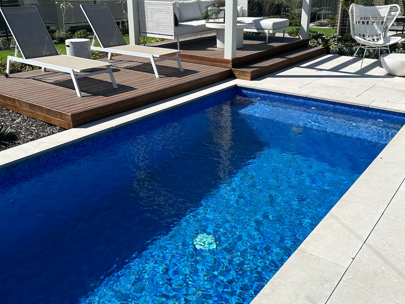 A blue tiled plunge pool with a broad shallow bench along one end, timber decking and sun lounges running down the long side.