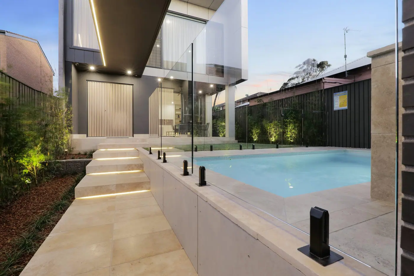 A plunge pool beside a contemporary home at dusk, with lit stone steps and paving leading down to the frameless glass fence.