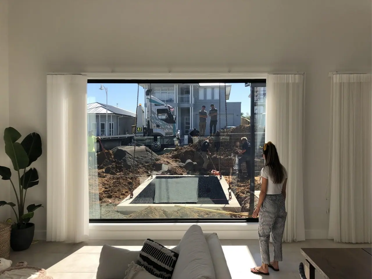 The view from inside a living room through a large window: the install crew and a truck outside, with the pool shell already set into the ground in the yard.